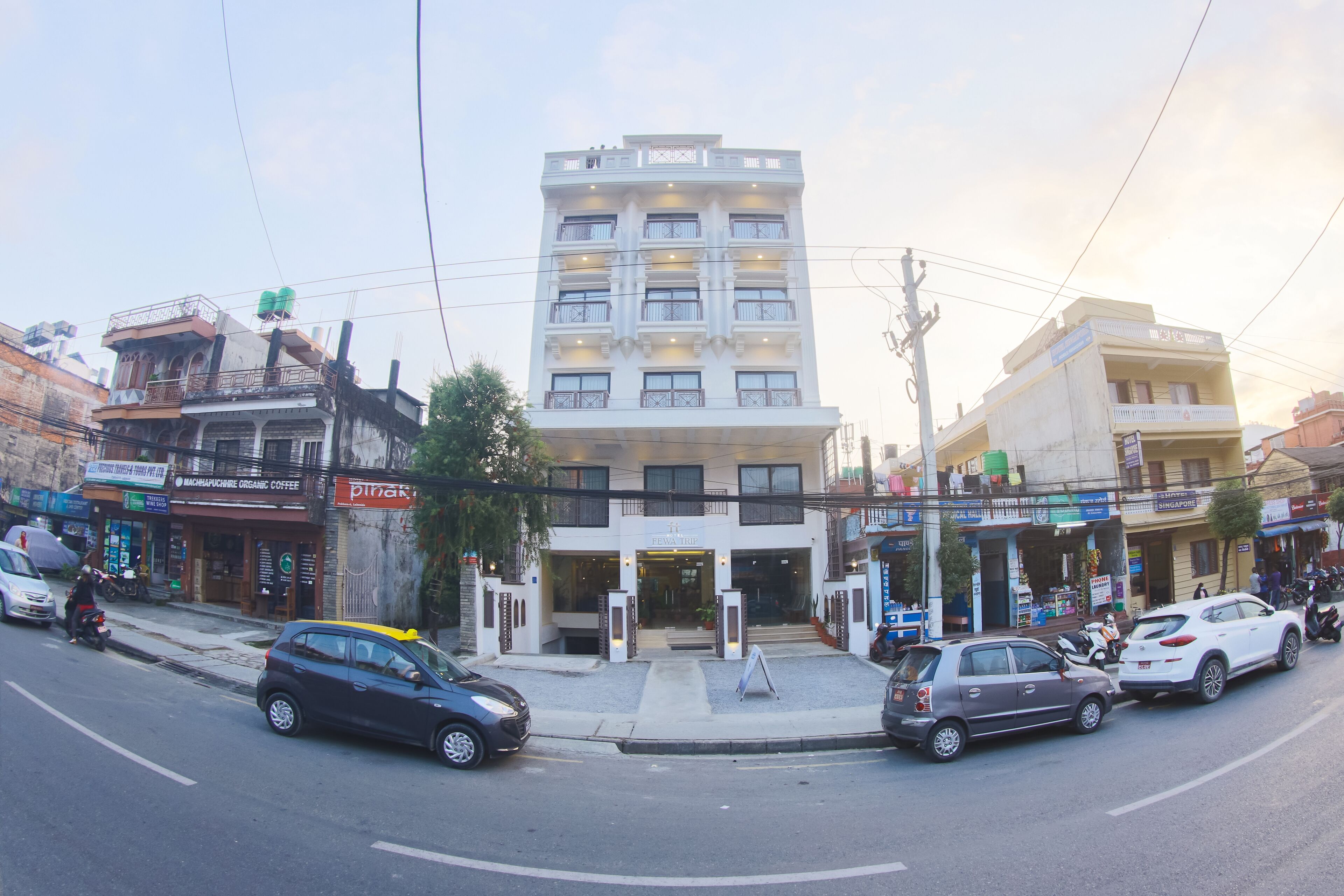 A multi-story hotel with a central entrance, surrounded by various shops and parked cars.