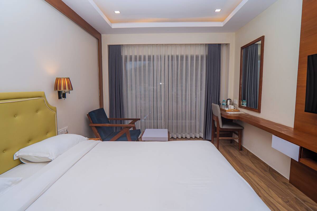 Habitación de hotel con cama, escritorio, silla, lámpara y un ventanal con cortinas.
