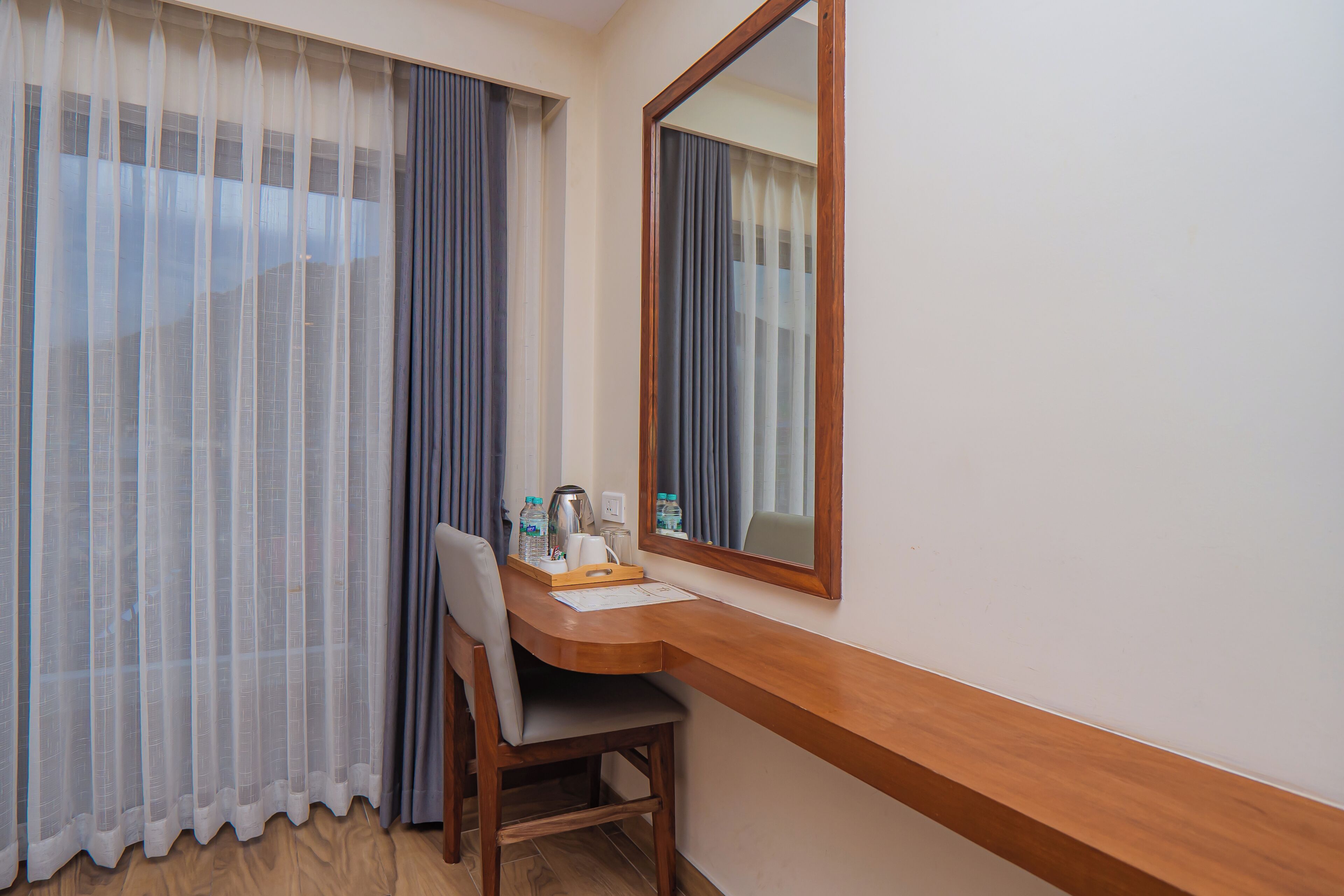 A hotel room with a wooden desk, a chair, a mirror, and a window with sheer curtains.