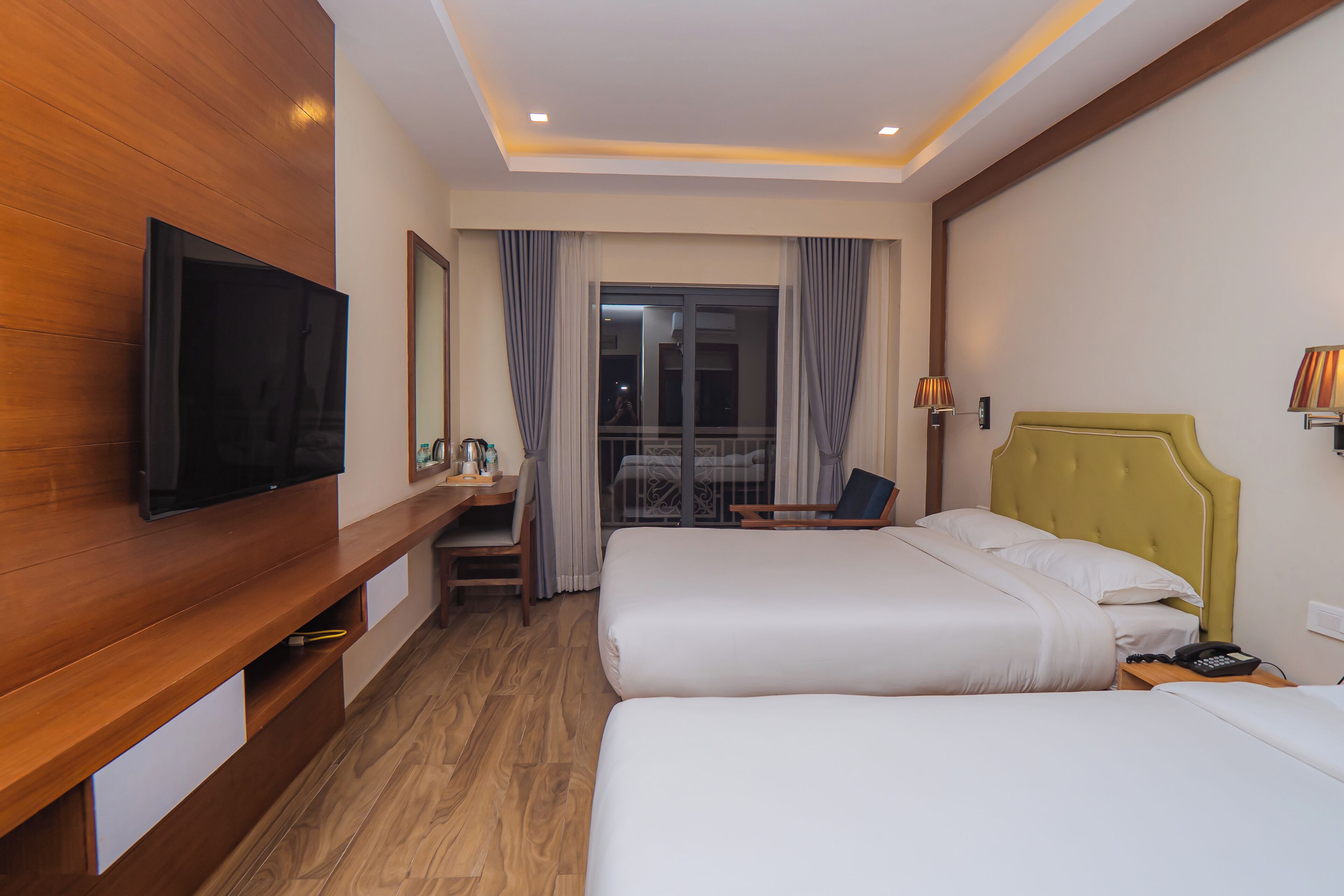 A hotel room with two beds, a TV, a desk, and a balcony with curtains.