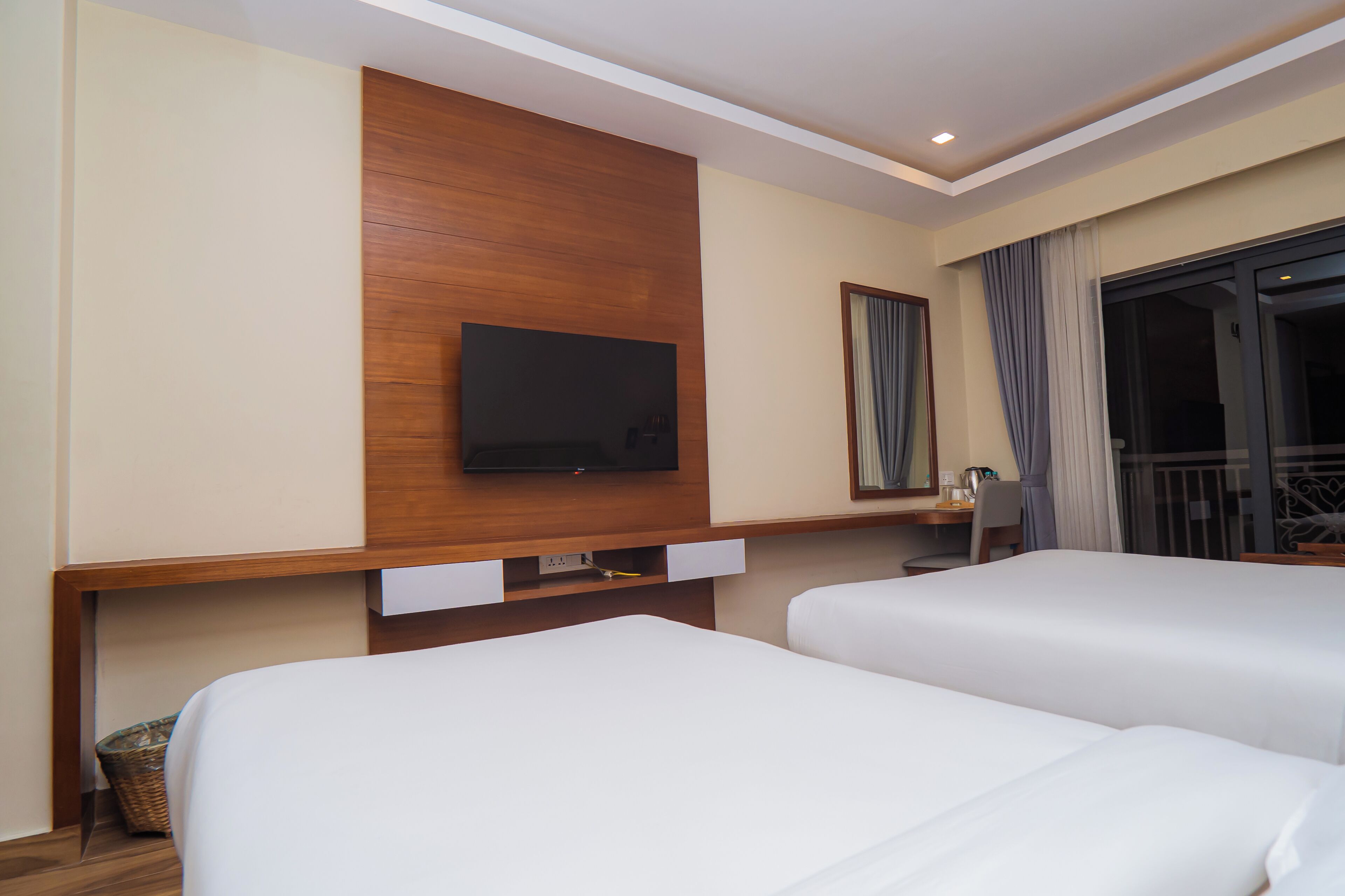 Habitación de hotel con dos camas, un televisor de pantalla plana montado en un panel de madera, un escritorio con una silla y una ventana con cortinas.