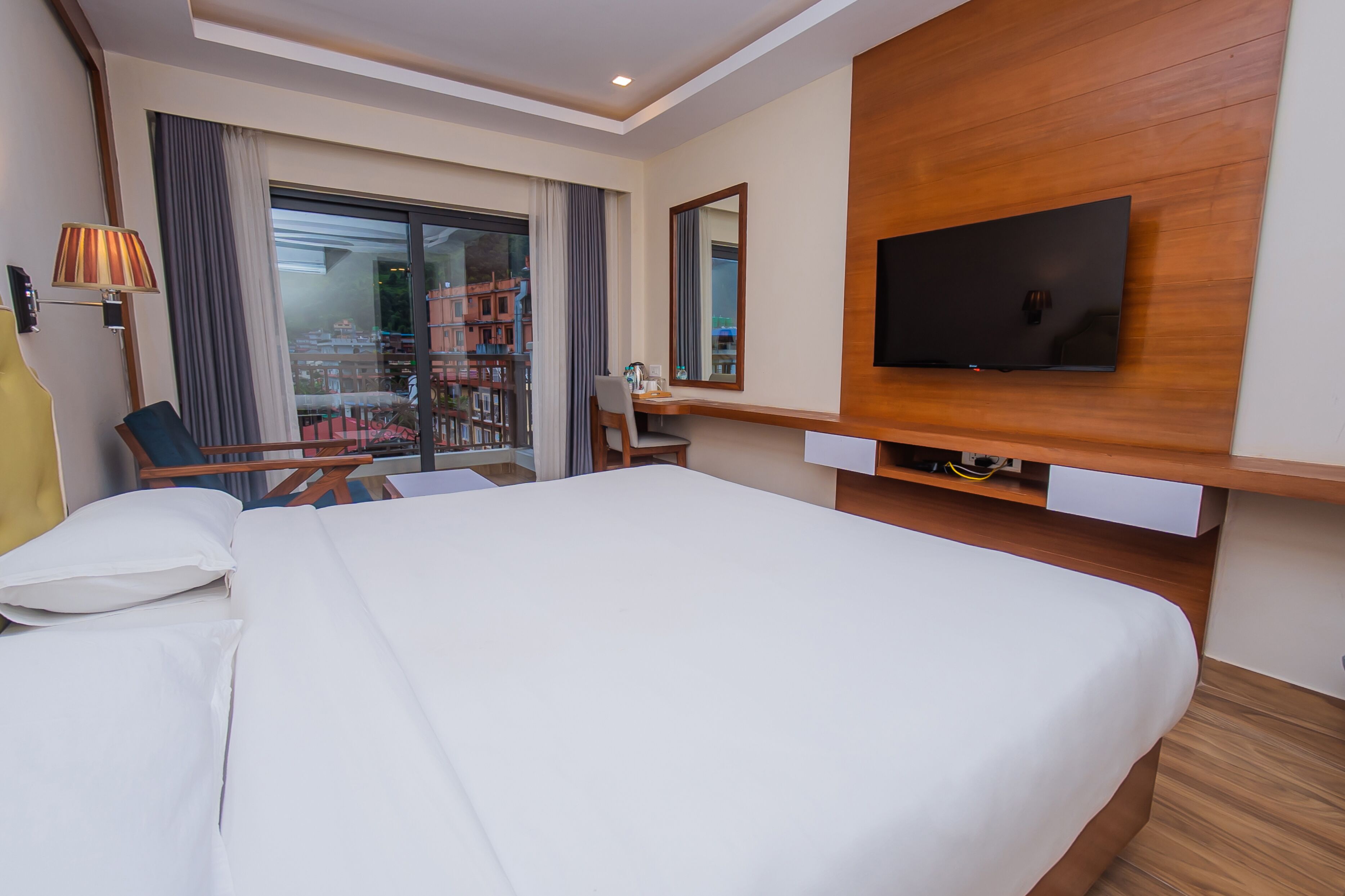 A hotel room with a large bed, a desk, a chair, a TV mounted on the wall, and a window with curtains.