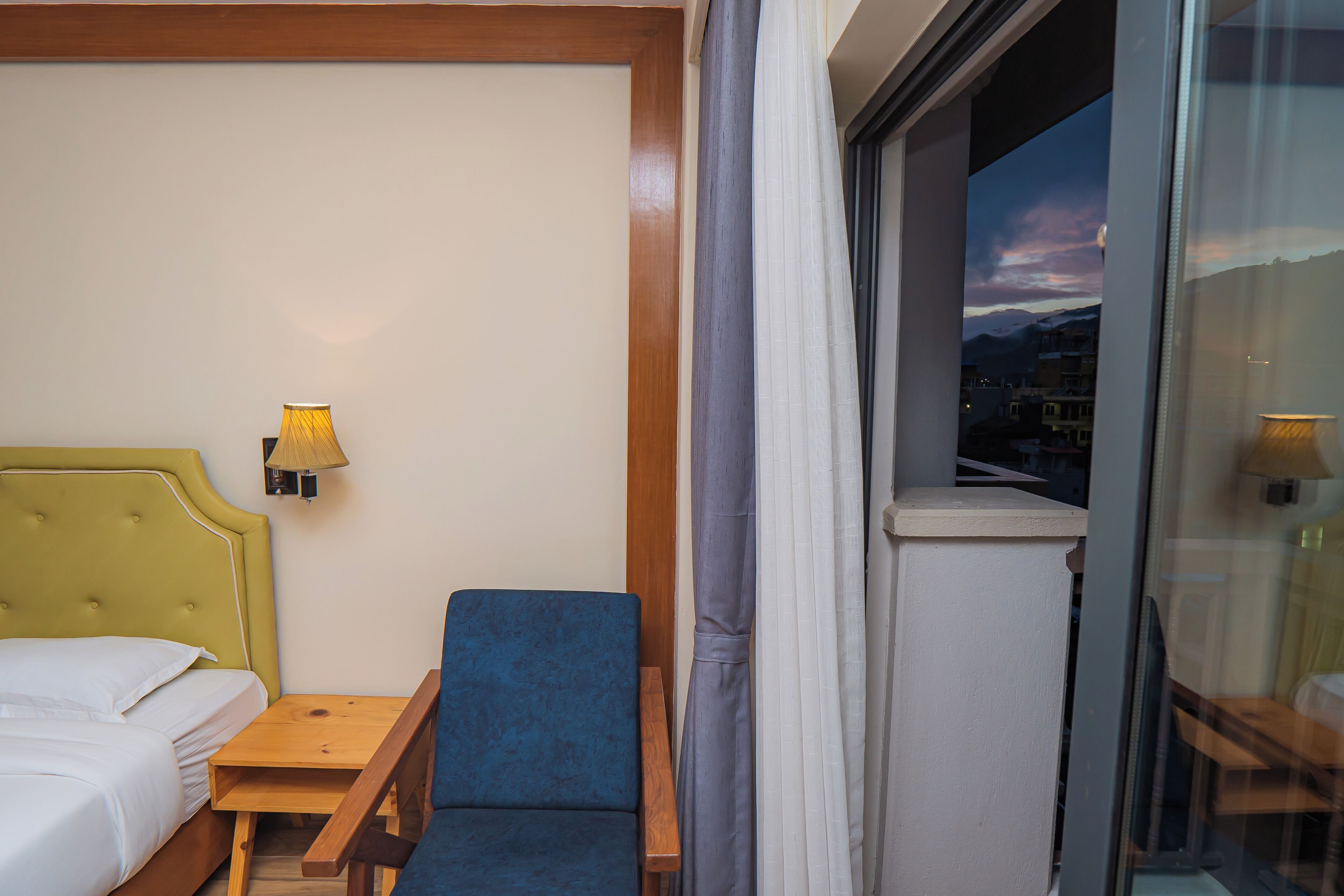 Habitación de hotel con cama, una silla azul, una mesita de noche de madera, una lámpara y vistas de un paisaje urbano a través de una ventana.