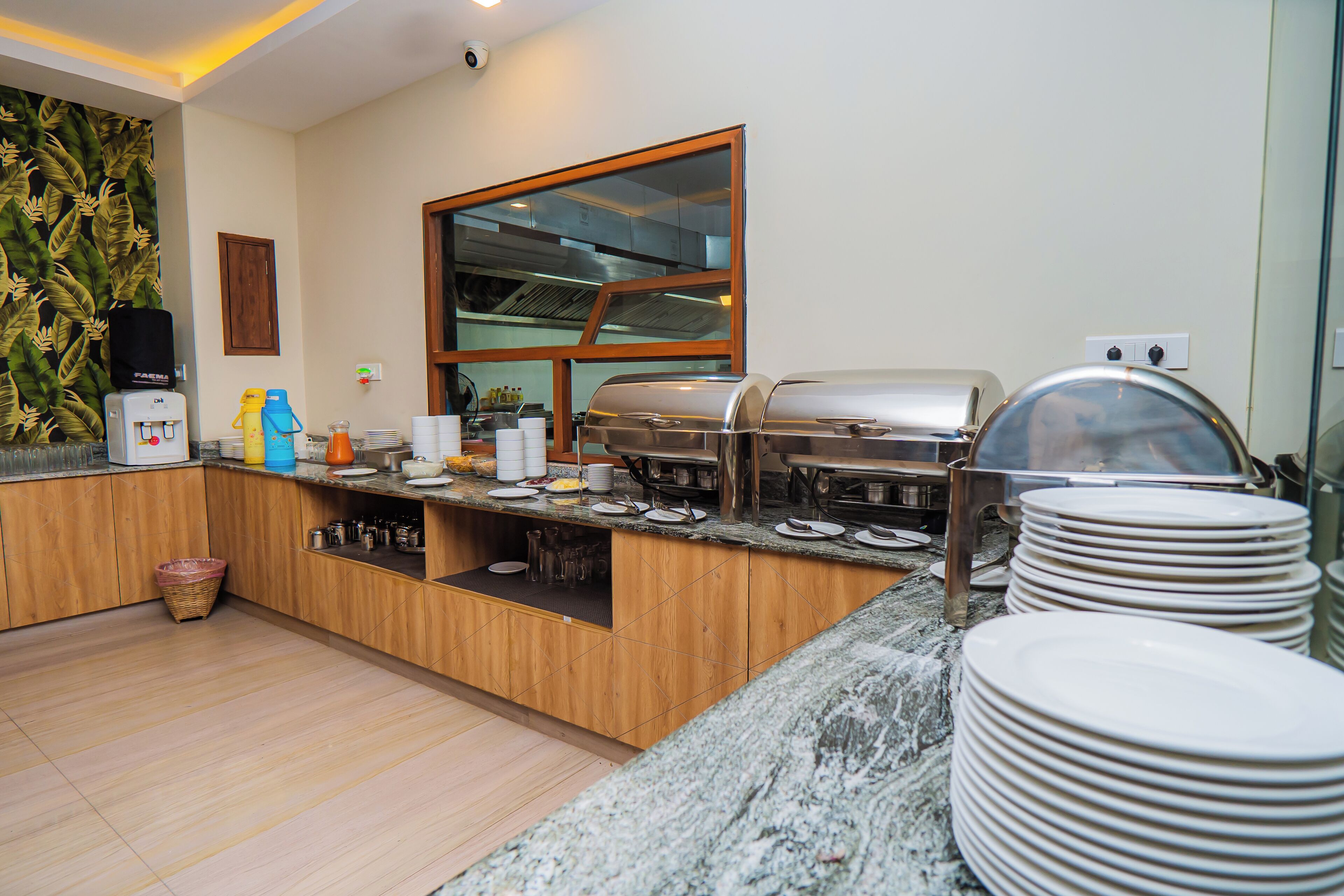 A breakfast bar with a water dispenser, coffee maker, and a grill. There are stacks of plates and a basket on the floor.
