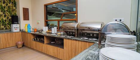 A breakfast bar with a water dispenser, coffee maker, and a grill. There are stacks of plates and a basket on the floor.