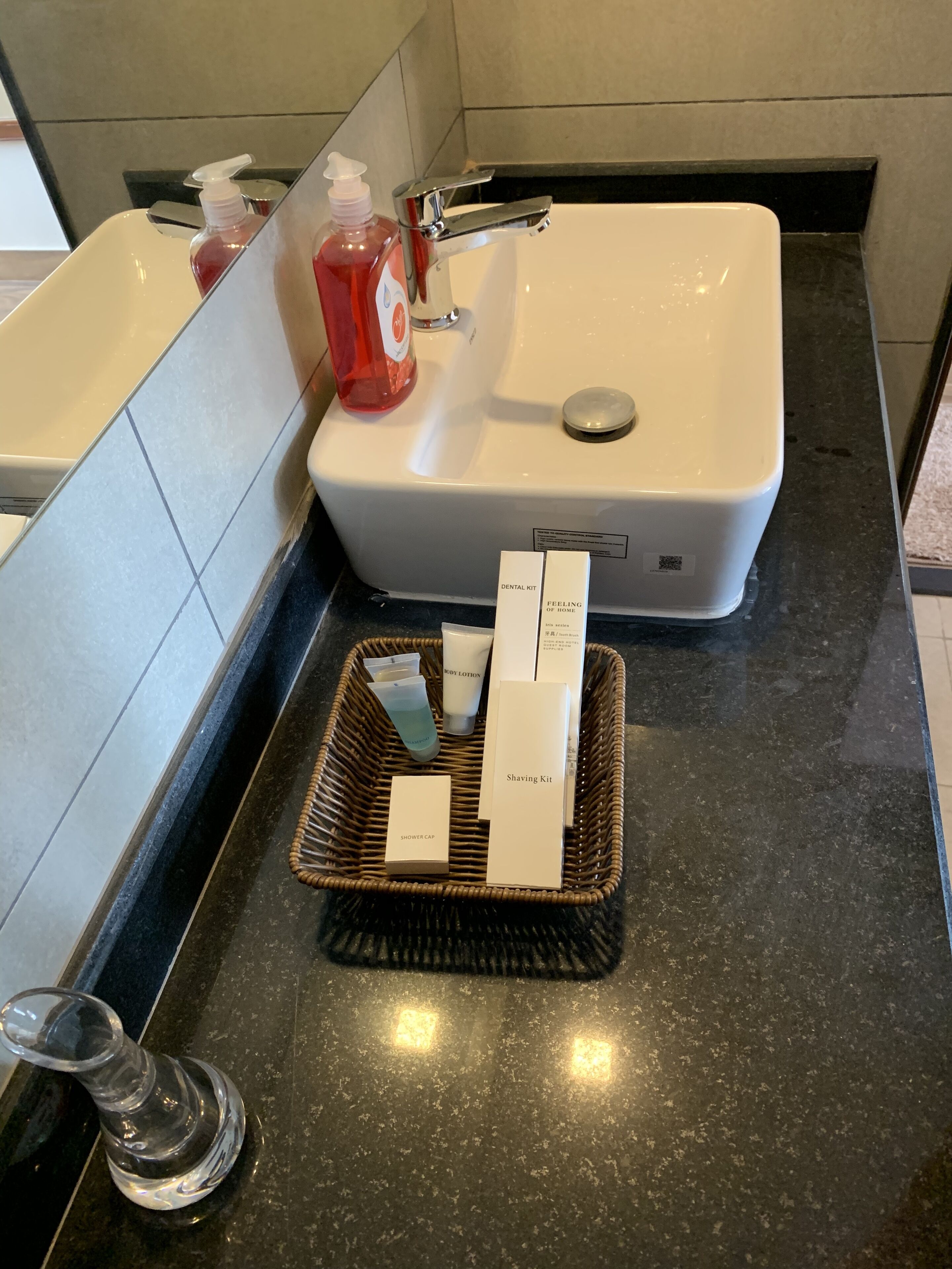 A bathroom with a white sink, a mirror, and a black countertop. There is a red bottle of soap and a white bottle of lotion on the sink. A wicker basket with a shaving kit and a shower cap is on the countertop.