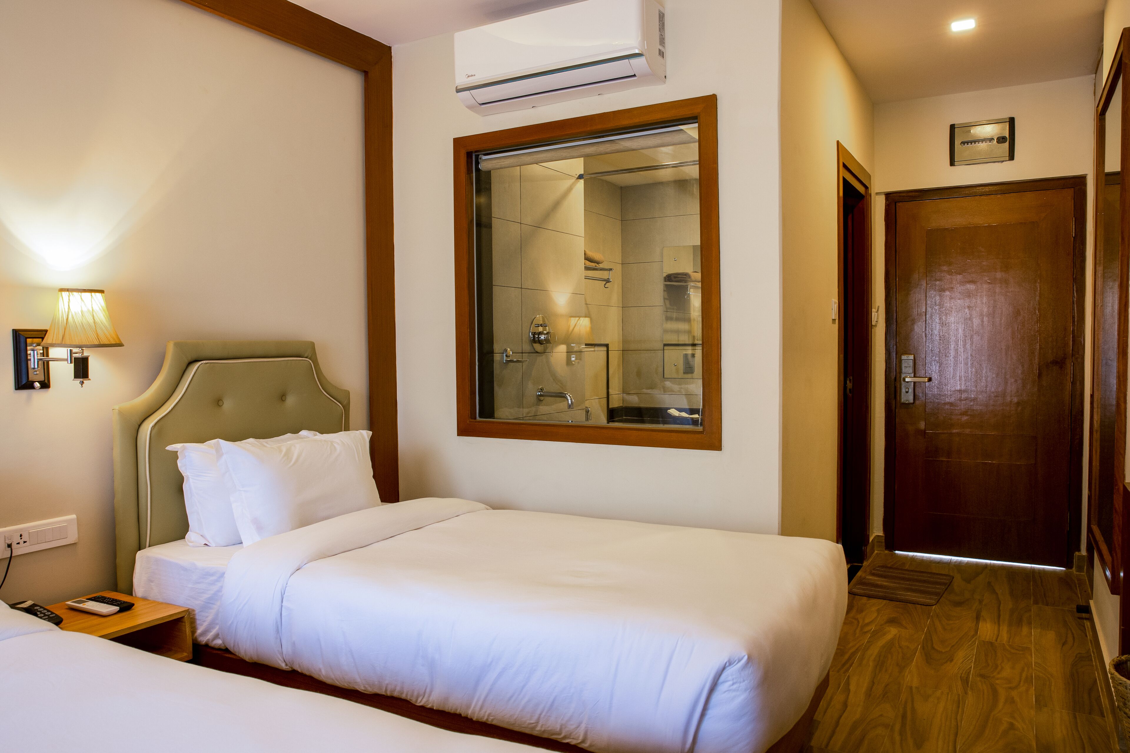 A hotel room with two beds, a nightstand, a lamp, a mirror, and a wooden door.
