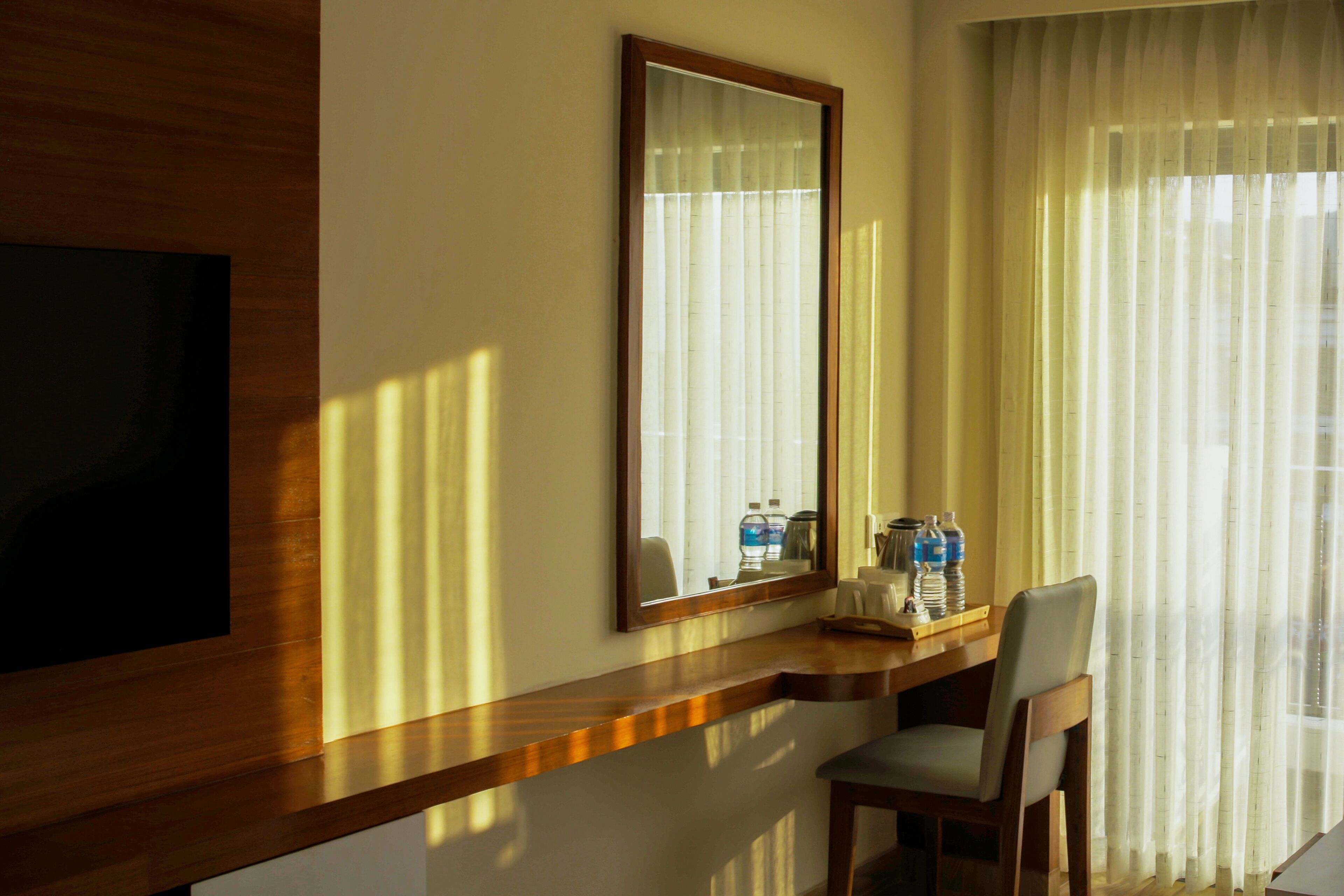 Habitación de hotel con un escritorio de madera, una silla, un espejo grande y una ventana con cortinas de encaje.