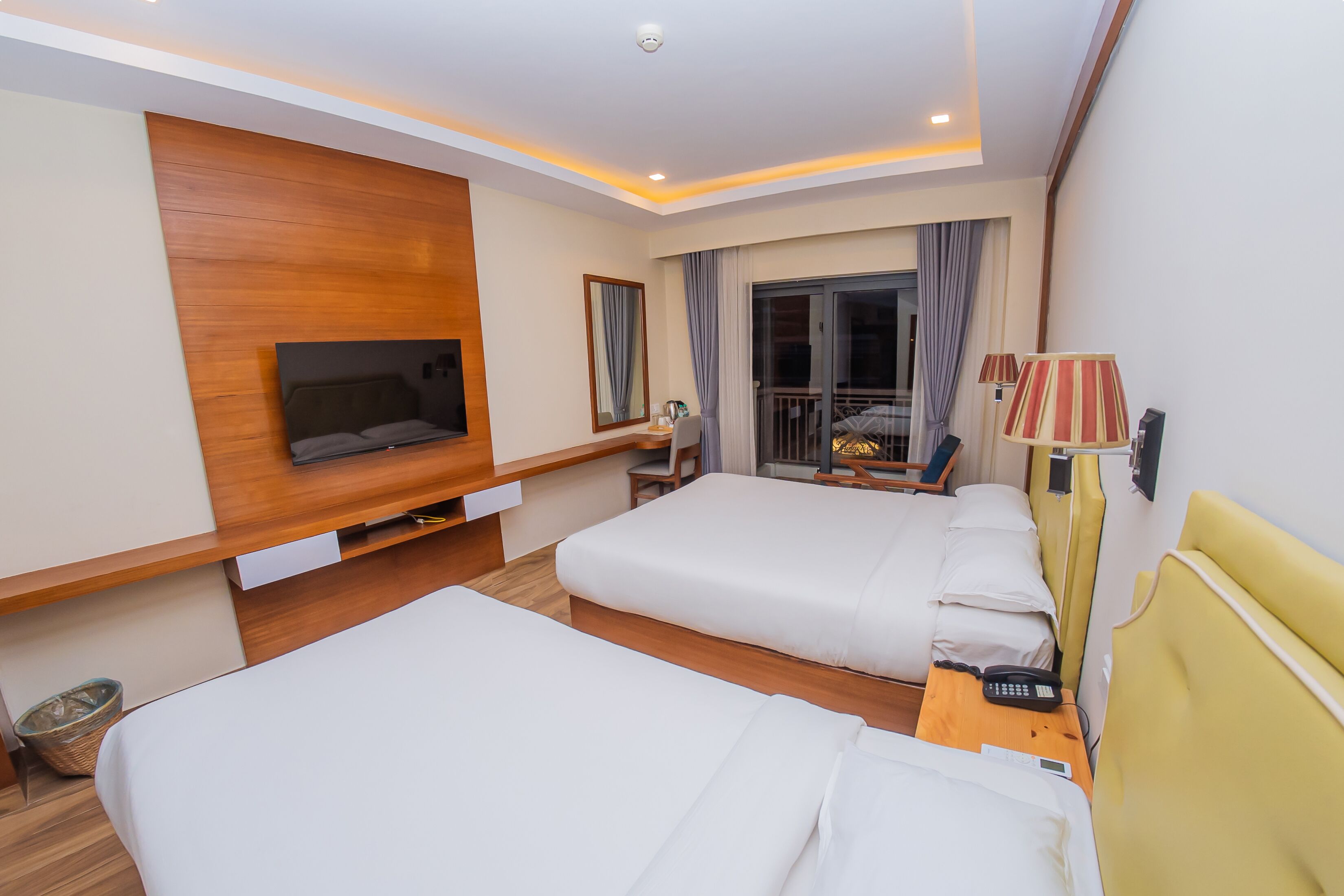 A hotel room with two beds, a TV, a desk, and a balcony with curtains.
