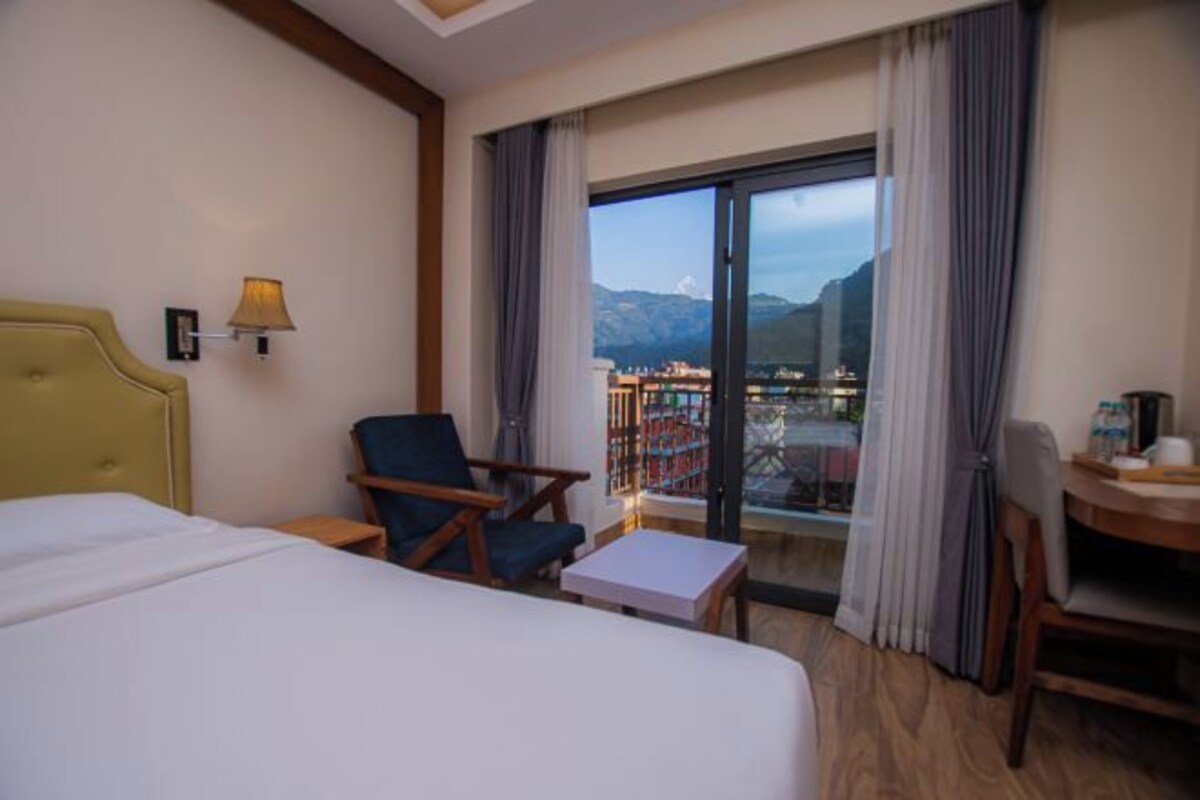 A hotel room with a large bed, a chair, a small table, and a view of a cityscape through a glass door.