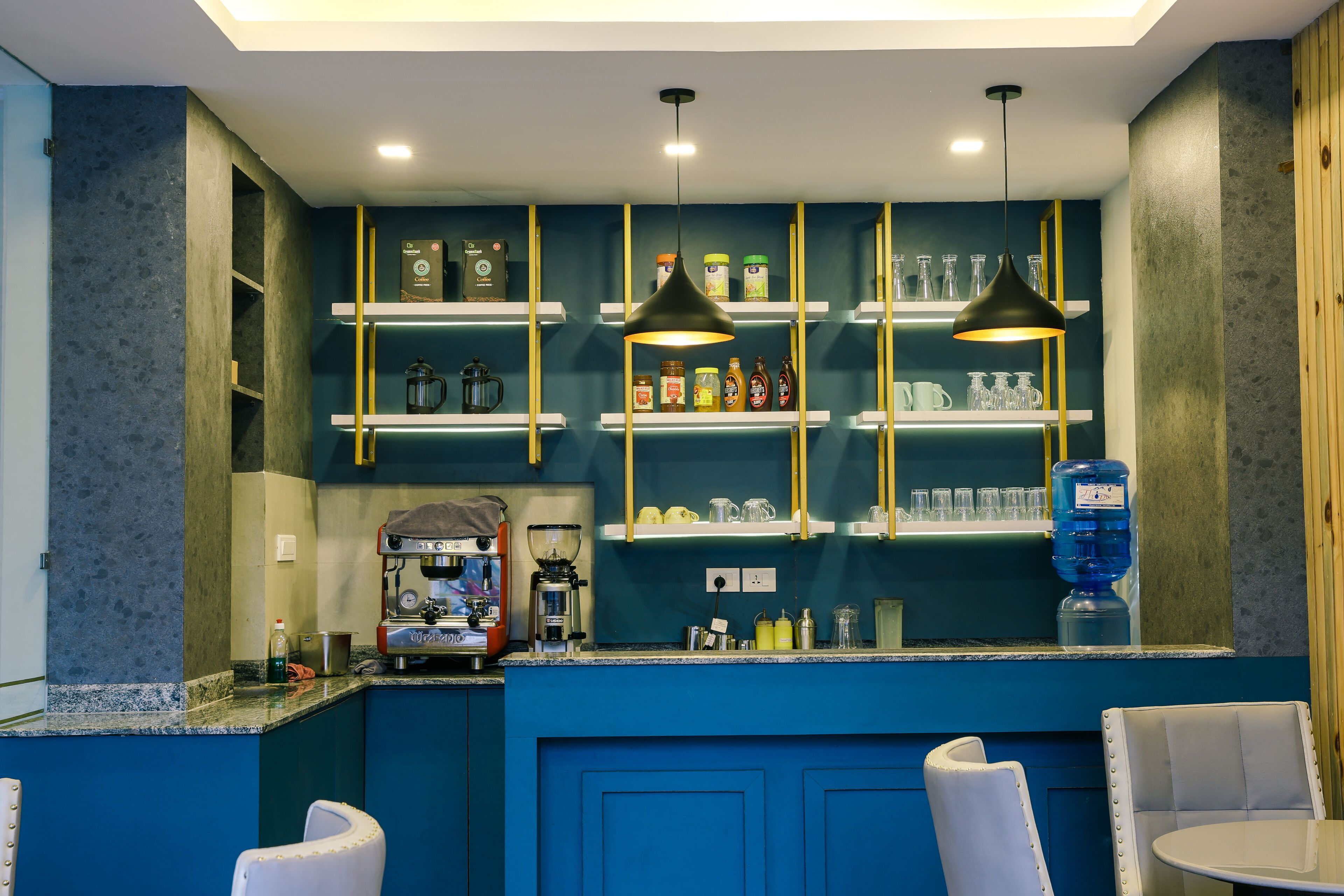 A bar area with blue cabinetry, shelves with bottles and glasses, and a coffee machine.