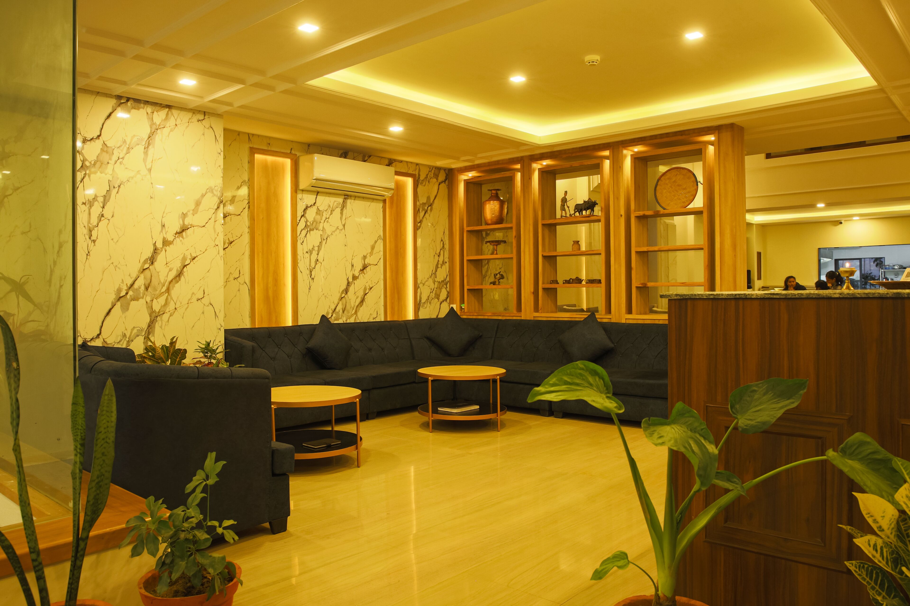 A modern lobby with a marble wall, dark sofas, wooden shelves, and potted plants.
