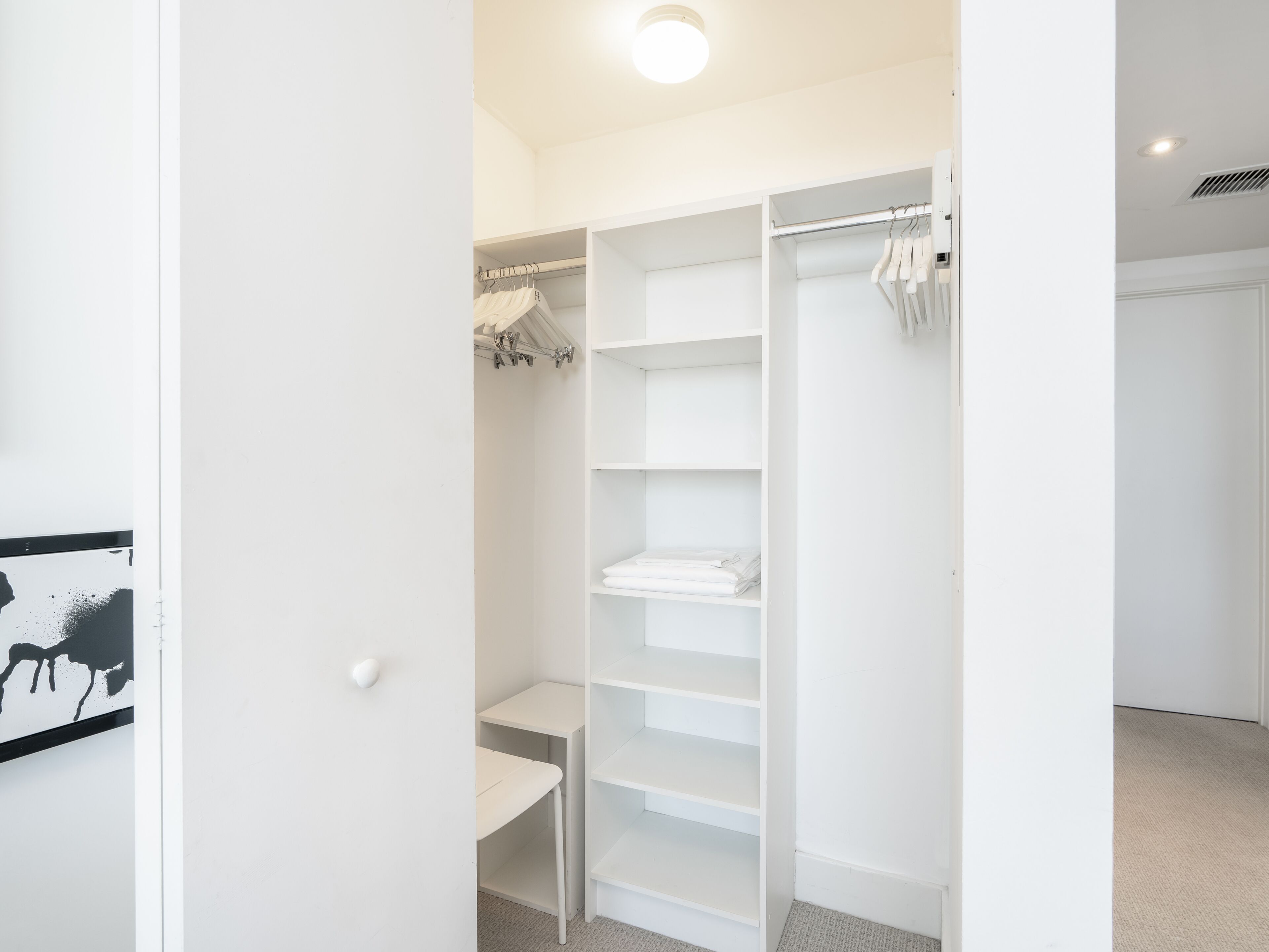 Un dressing ouvert de couleur blanche, doté d’étagères et d’un espace pour suspendre des vêtements, une petite table et une porte donnant sur une autre pièce.