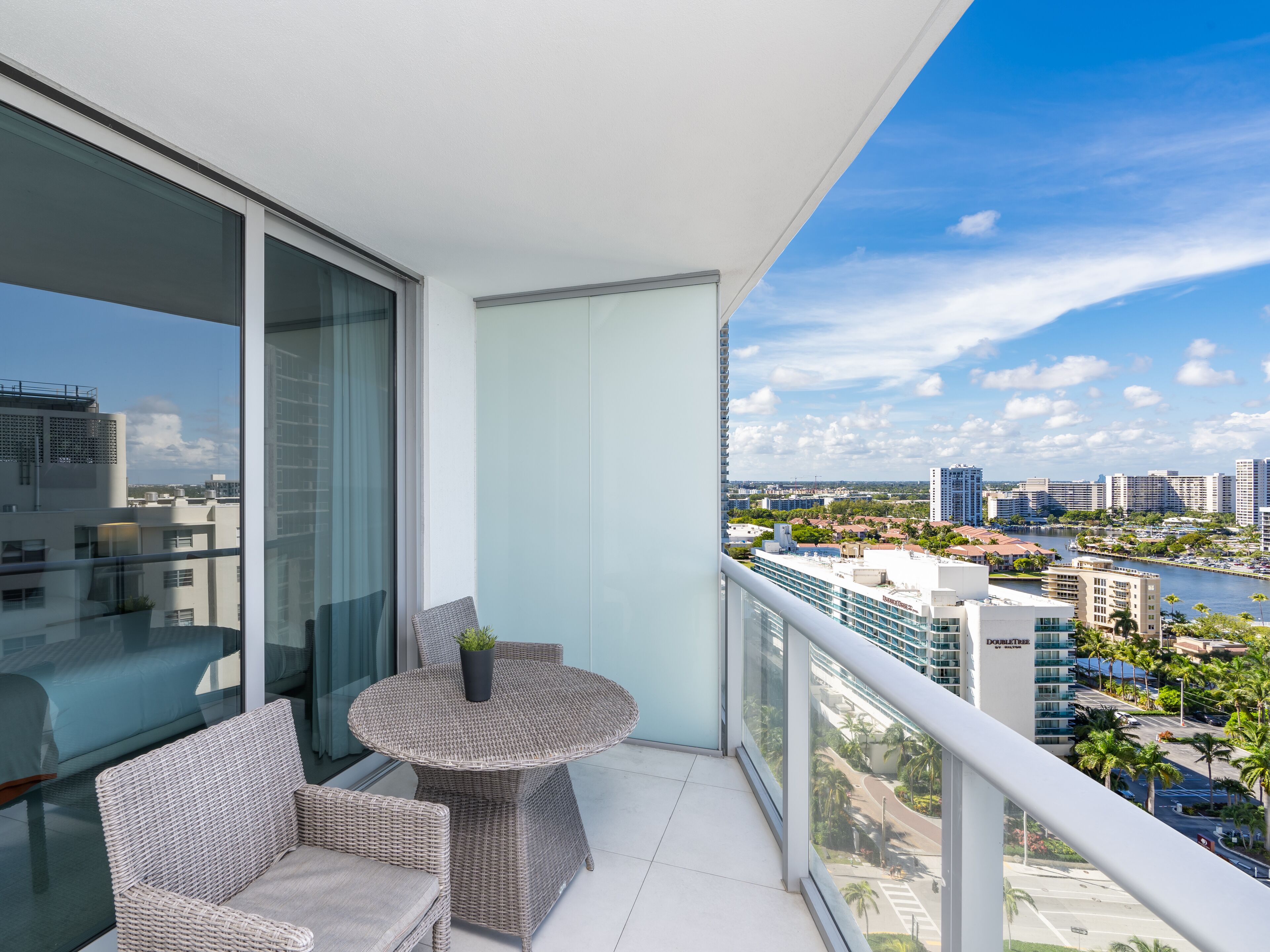 A balcony with wicker furniture, a glass table, and a view of a cityscape and water.