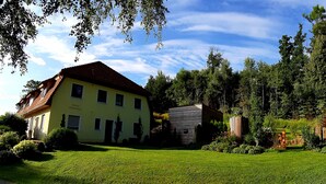 Une maison jaune avec un toit rouge, entourée d’un jardin bien entretenu et de grands arbres.