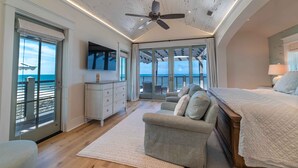 A bedroom with a large bed, a ceiling fan, a TV, and a view of the sea through the windows. - Luxury beach-front home, gourmet kitchen, private pool, gated community, gear (Port Saint Joe)