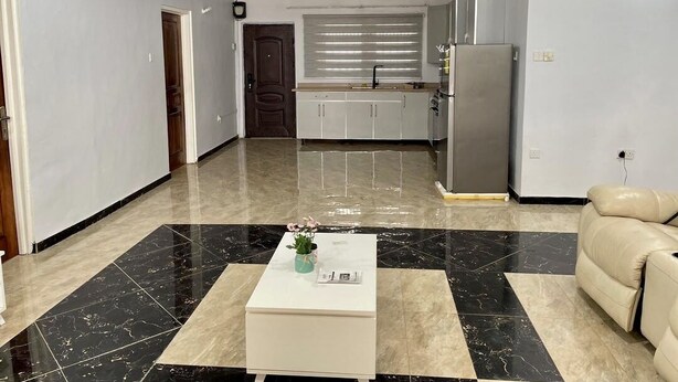 Un espacio habitable amplio, con una cocina al fondo, una mesa de centro blanca y un sofá color beige.