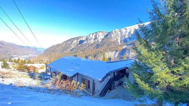 Un bâtiment enneigé, doté d’un escalier menant à un deuxième étage, entouré de montagnes et d’arbres.