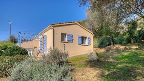A single-story house with a beige exterior, two windows with blue shutters, a blue metal staircase, and a garden with various plants and a large rock. - L' Amandier with communal swimming pool (AJACCIO)