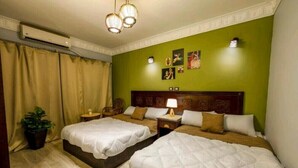 Quarto de hotel com duas camas, uma cabeceira de madeira, uma mesa de cabeceira com abajur e um vaso de planta.