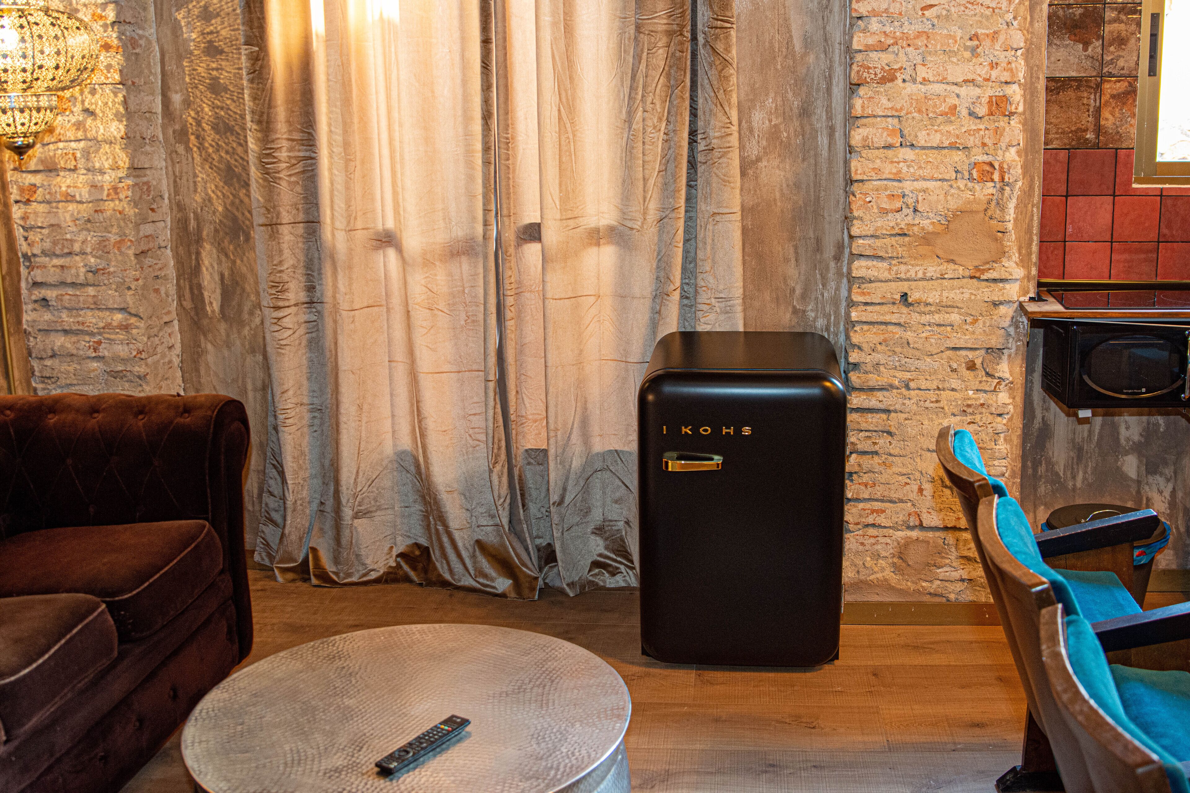 Una habitación acogedora con un sofá antiguo, una mesita redonda con un control remoto, un refrigerador IKOHS negro y una silla de madera con un cojín azul.