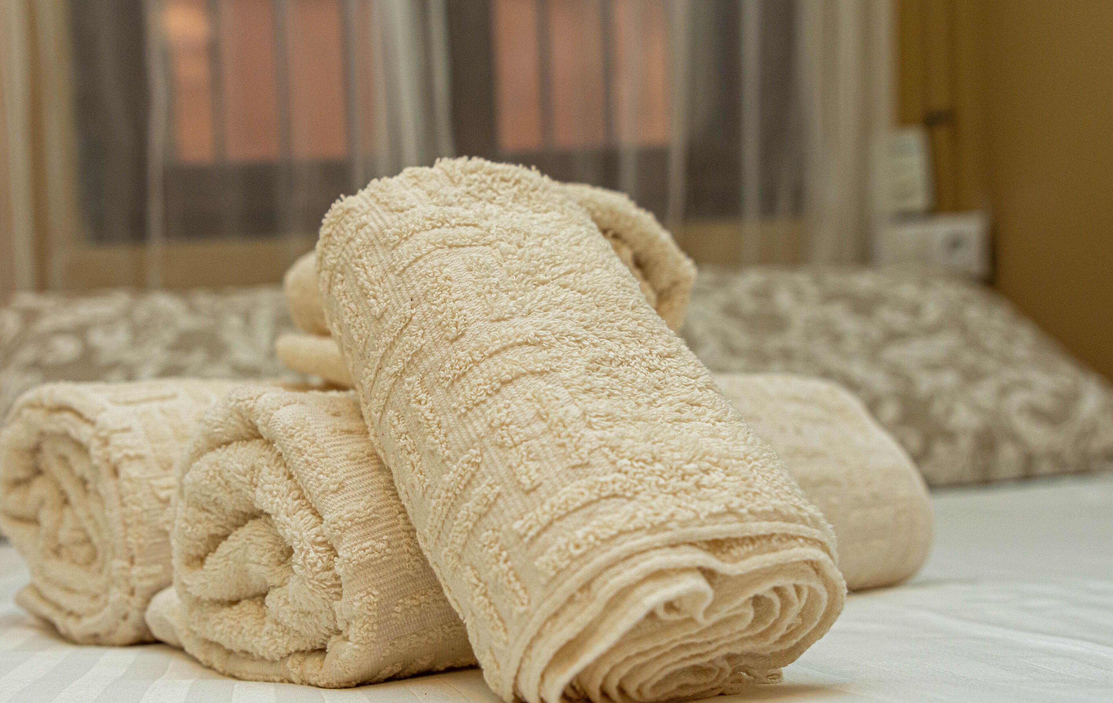 En prydligt vikt, beige handduk ligger på en säng, och i bakgrunden syns en kudde med ett mönster.
