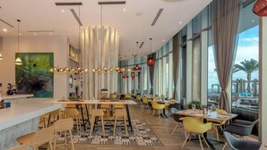 Un ristorante moderno con un lungo bancone, diversi tavoli e sedie. Grandi finestre offrono vista all'esterno.