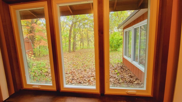 A view through a set of three windows, revealing a backyard with a wooden deck, a shed, and a forested area with autumn leaves. - Relax at this Private, Peaceful, Dog-Friendly Ecohouse in the Woods. (Lowell)