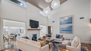 Ein geräumiges Wohnzimmer mit hoher Decke, weißen Wänden und einem großen Fenster. Zwei weiße Sofas, ein Couchtisch und ein Essbereich mit Korbstühlen sind vorhanden.