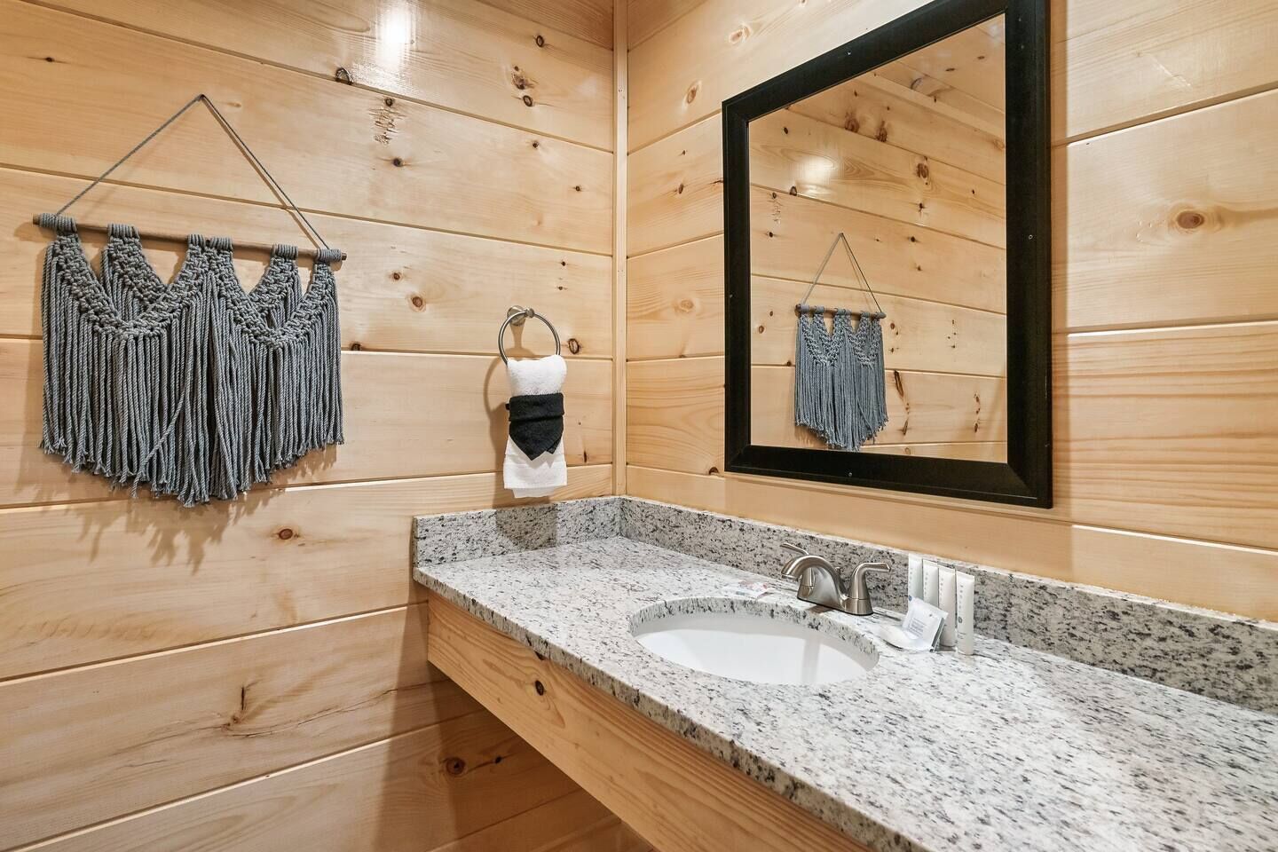 Un bagno in stile baita con elementi in legno, un lavabo con piano in granito, un porta asciugamani sospeso con asciugamani in macramè e uno specchio con cornice nera.