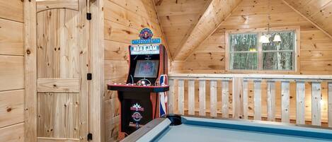 Eine Holzhütte mit Billardtisch, NBA Jam Spielautomat und einem Fenster mit Blick auf Bäume.