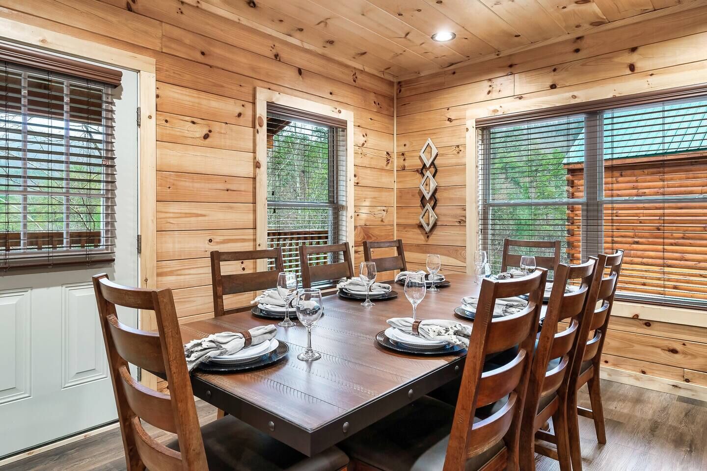 A dining area with a wooden table set for six, surrounded by chairs, with large windows and a view of greenery outside.