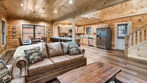 A cabin interior with a wooden floor, a leather sofa, a dining table, and a kitchen area. - Best Location 5BR Sleeps 16 (Pigeon Forge)