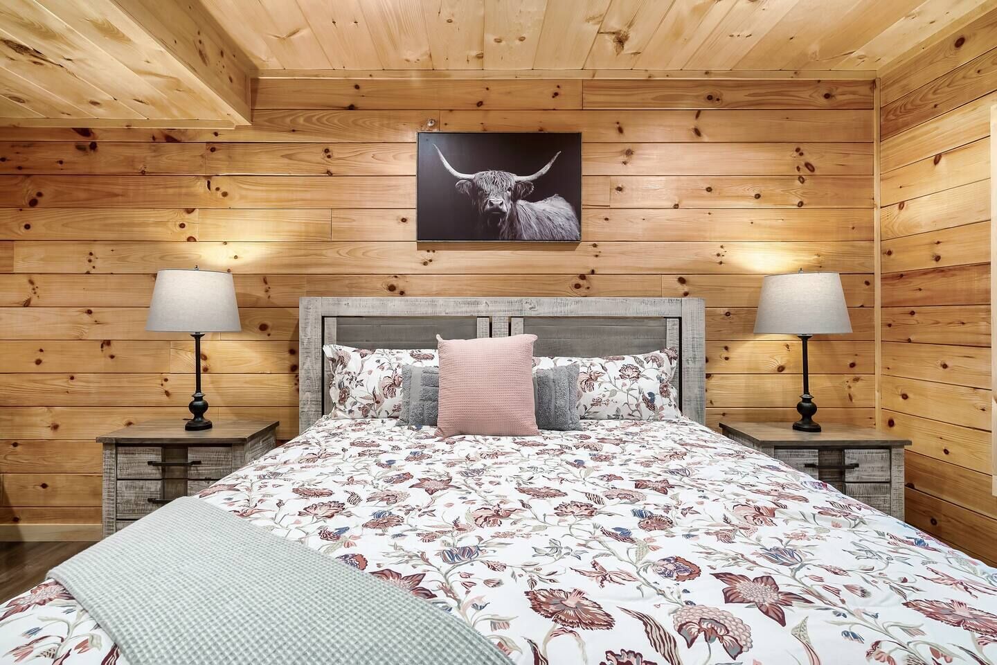 A wooden-paneled bedroom with a bed, two bedside lamps, a floral bedspread, and a picture of a bull's head on the wall.