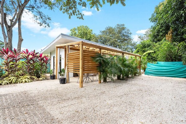 A modern outdoor structure with a wooden pergola, surrounded by gravel, potted plants, and trees.