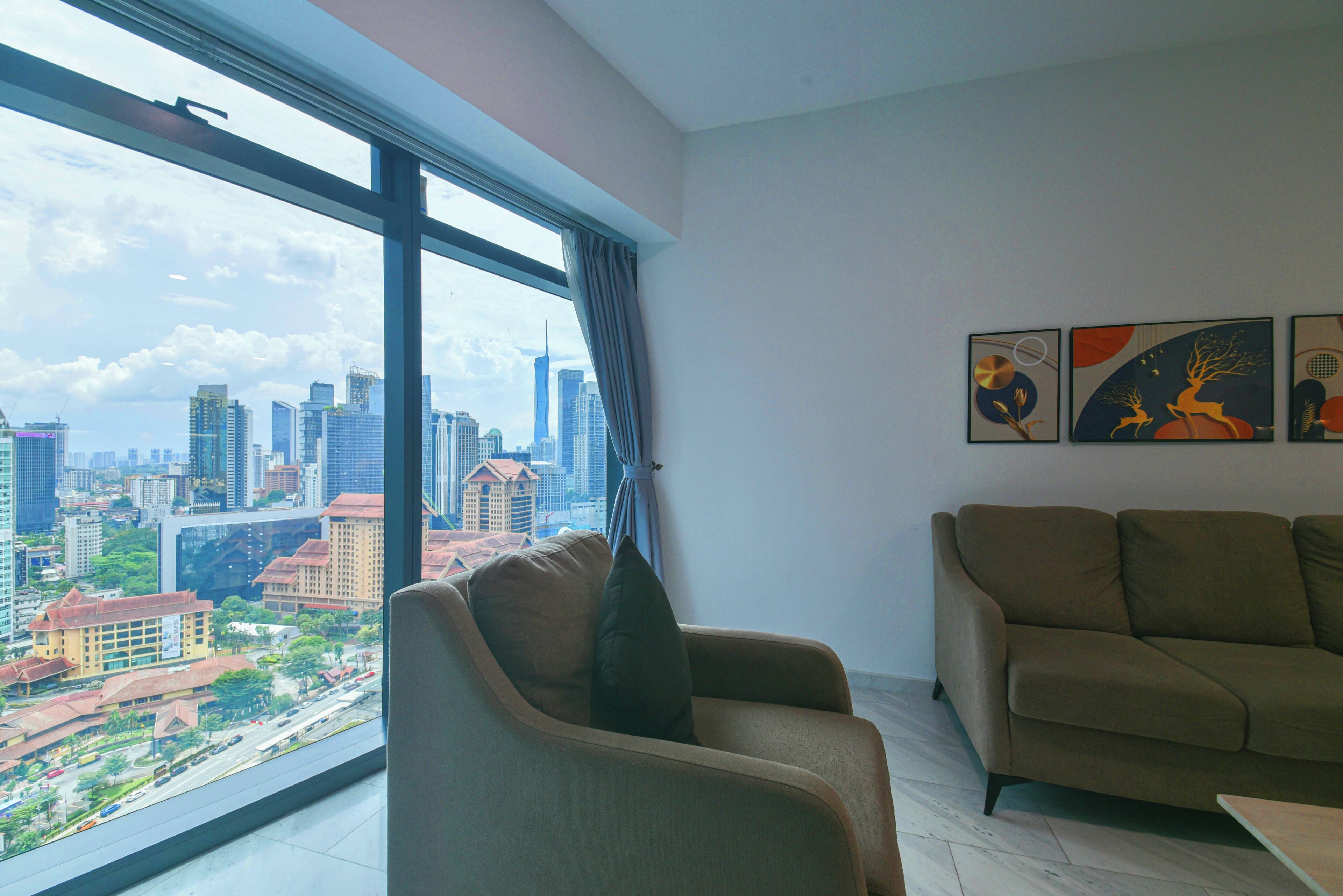A modern living room with a large window offering a city view, a beige sofa, and three framed artworks on the wall.