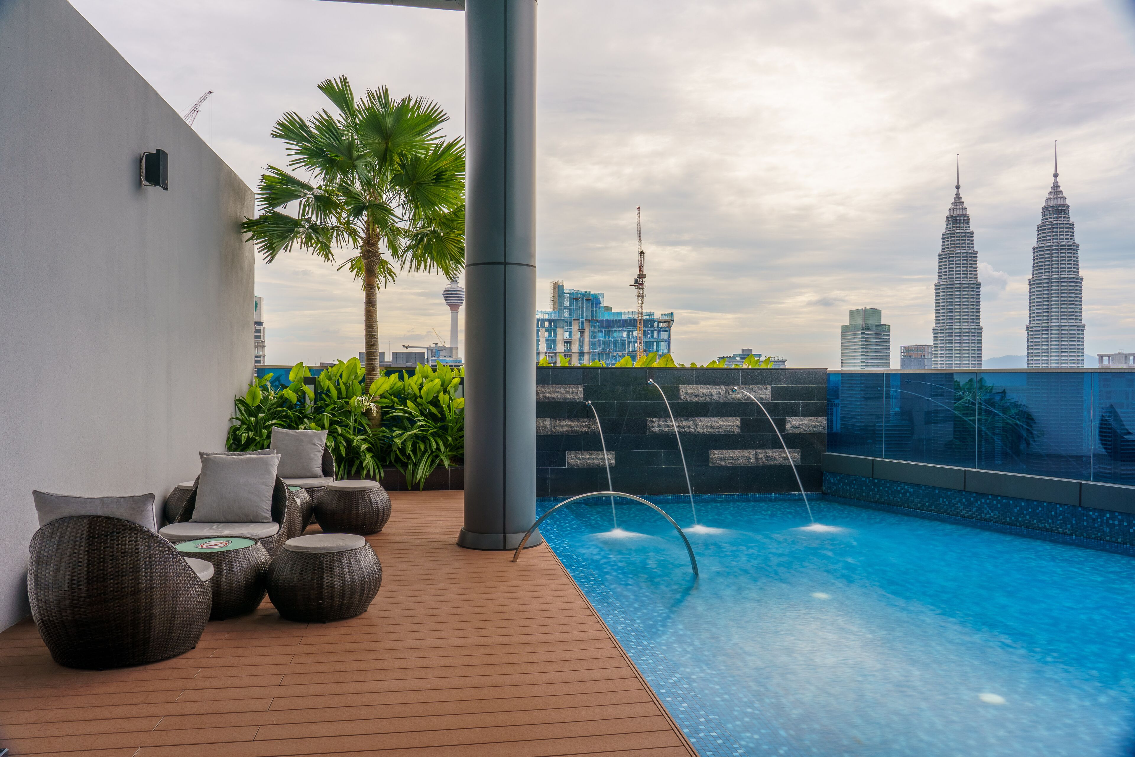 A rooftop pool with a view of the Petronas Towers, surrounded by wicker furniture and palm trees.