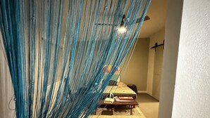 A room with a blue hanging fabric, a bed with a striped blanket, and a small table with a lamp. - Remodeled quiet and spacious home. (Blythe)