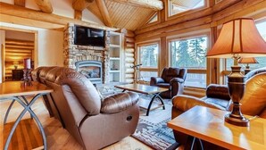A log cabin interior with a stone fireplace, leather seating, and large windows.