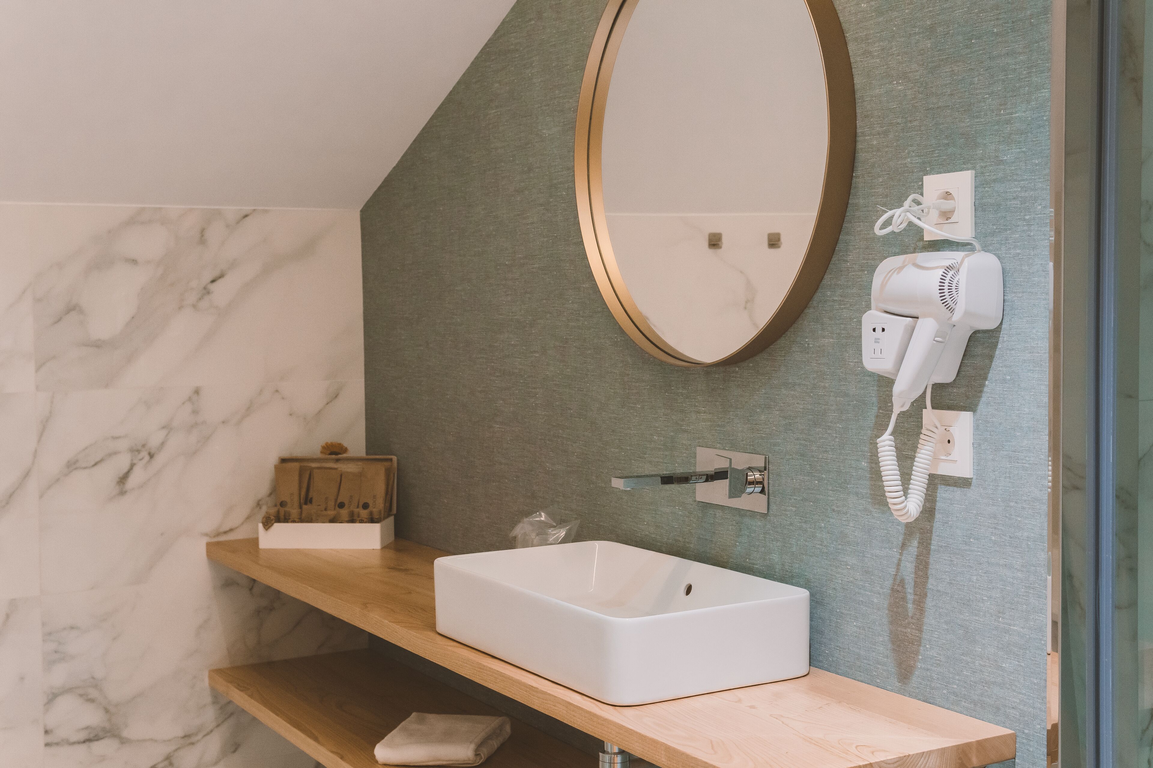 A bathroom with a marble wall, a wooden vanity, a white sink, a round mirror, and a hairdryer on the wall.