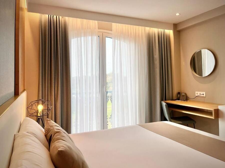 A hotel room with a bed, a desk, and a chair. There is a window with curtains and a view of the outdoors.