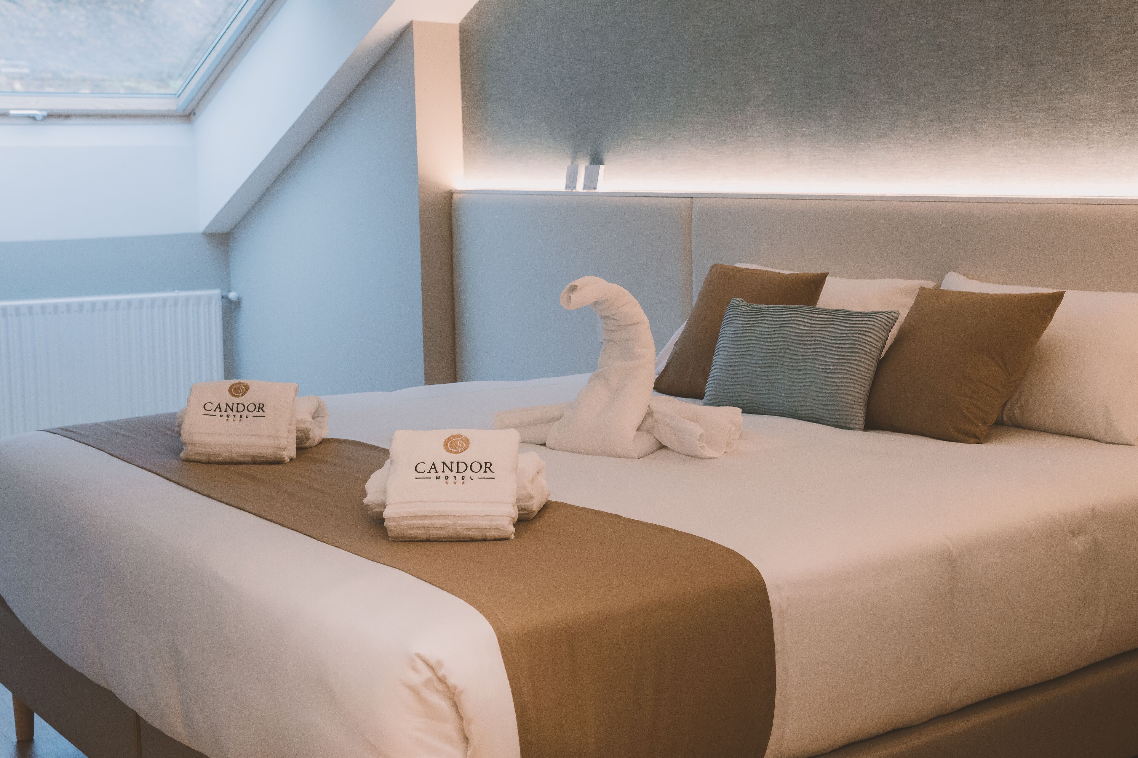 Ein Hotelzimmer mit einem ordentlich bezogenen Bett, einem Kopfteil und Handtüchern mit dem Hotelnamen.