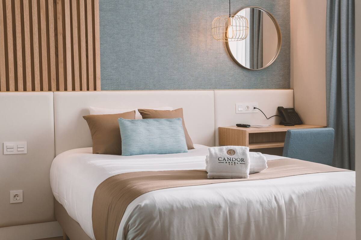 Ett hotellrum med en sÀng, ett nattduksbord med en telefon, en lampa och en handduk med hotellets namn pÄ.