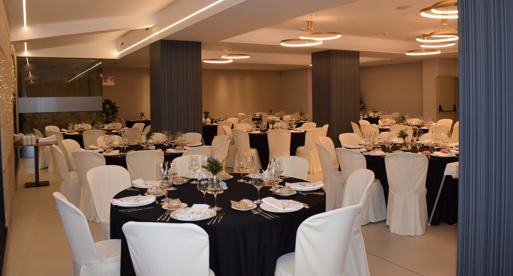 A banquet hall with round tables set for dining, white chairs, and modern lighting fixtures.