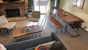 A living room with a fireplace, a TV mounted above it, a dining table with benches, a sofa, and a large window with curtains. - Ski condo close to multiple resorts (Mendon)