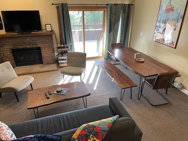 Ein Wohnzimmer mit Kamin, einem darüber hängenden Fernseher, einem Esstisch mit Bänken, einer Couch und einem großen Fenster mit Vorhängen.