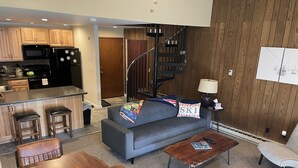A modern living room with a gray sofa, wooden coffee table, and a dining table. A kitchen with a black refrigerator and microwave is visible in the background. - Ski condo close to multiple resorts (Mendon)