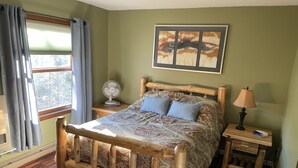 A wooden bed with a patterned bedspread, a bedside table with a lamp, a wall-mounted artwork, and a window with curtains. - Ski condo close to multiple resorts (Mendon)