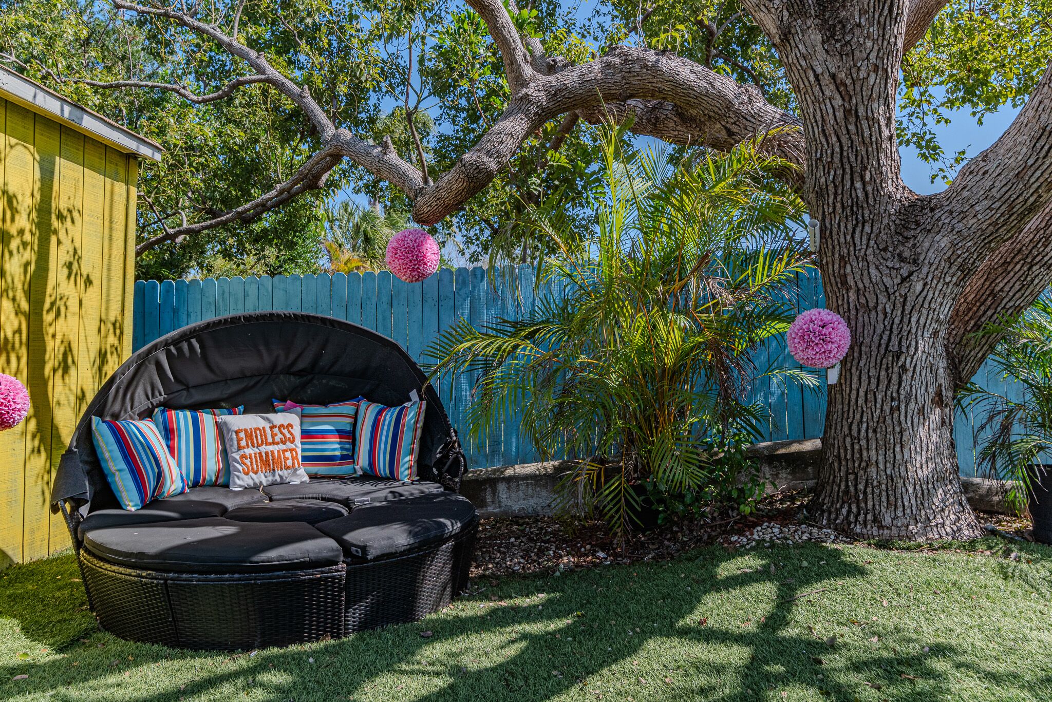 A black outdoor seating area with striped cushions and a pillow labeled "ENDLESS SUMMER," surrounded by a blue fence and lush greenery.