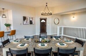 Une salle à manger avec une table en bois dressée pour six personnes, entourée de chaises. Un lustre se trouve au-dessus de la table, et un escalier avec une rampe est visible au fond.